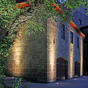 Mit dem Beleuchtungssystem rinnea wird ein altes Backsteingebäude in Szene gesetzt. Die LED Lichtstrahler im Boden strahlen die Hauswand bis unter das rote Dach an.