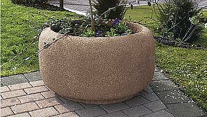 Das Pflanzengefäß Florenz in der Farbe rinnit Rotgranit steht vor einer grünfläche und ist bepflanzt mit einem Margeriten Strauch.