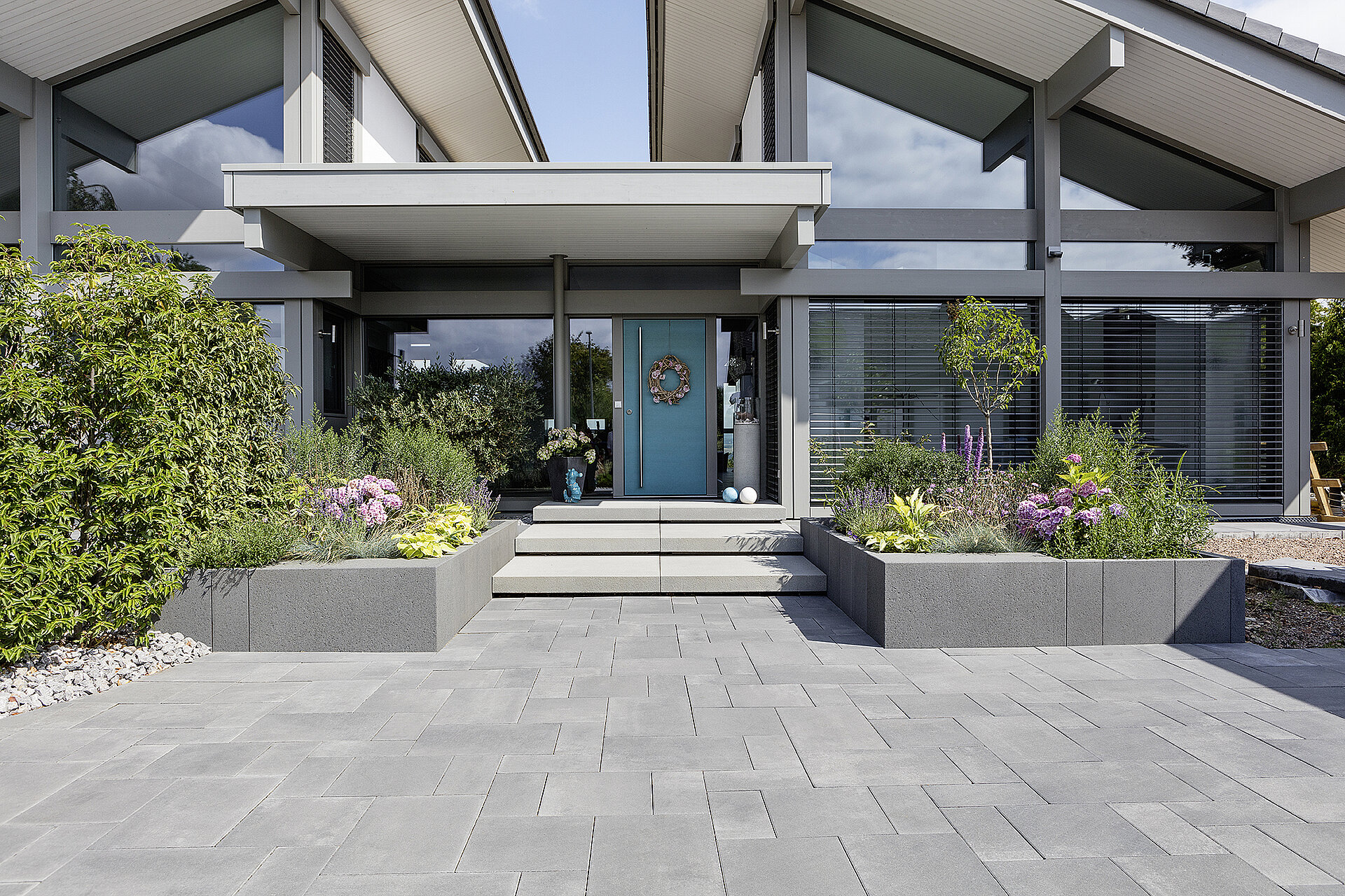 Beispielbild der Pflasterstein Farbe: Ein Eingang zum modernen Neubau mit zwei Pflanzbeeten links und rechts, türkisfarbener Haustür und wild verlegten Terrassenplatten in dunkelgrau.