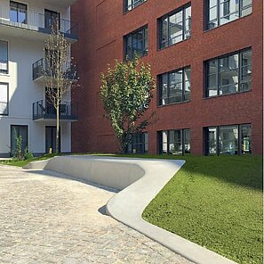 Vor einem Wohnhaus mit weißer Fassade auf der einen Seite und einer Backsteinfassade auf der anderen Seite ist ein Platz. Dort führt ein Weg entlang einer Grünfläche. Diese werden durch ein hellgraues Betonteil getrennt, welches als Sitzbank dient. Der Weg ist mit hellgrauen Pflastersteinen verlegt.